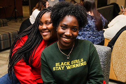 Two students of color smiling, one with a shirt reading Black Girls Stay Winning