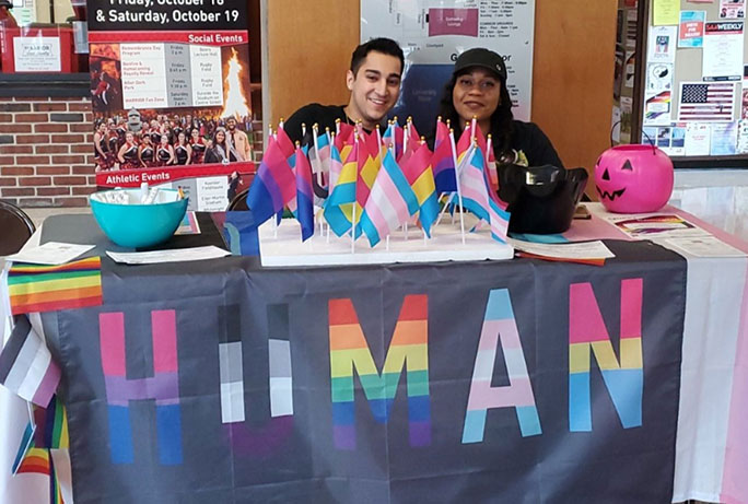 Two students behind a table with several LGBTQ+ flags and the word "Human"