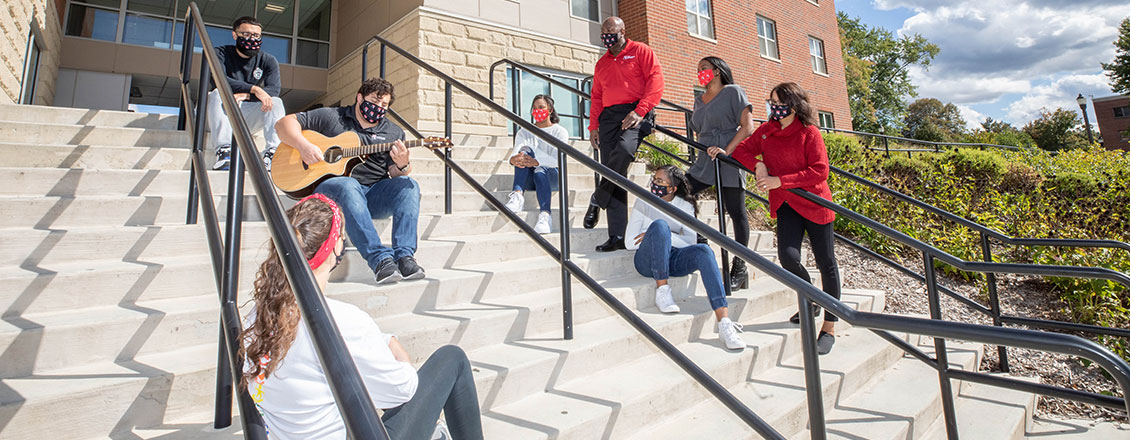 Interim President Kenneth Long standing in front of Sycamore Suites with students, one playing the guitar.