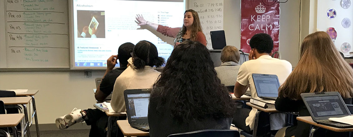 A secondary education teaching a class and gesturing to information displayed on a screen