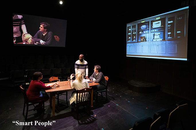 Actors sit around a table on a dark stage, with images projecting on two walls